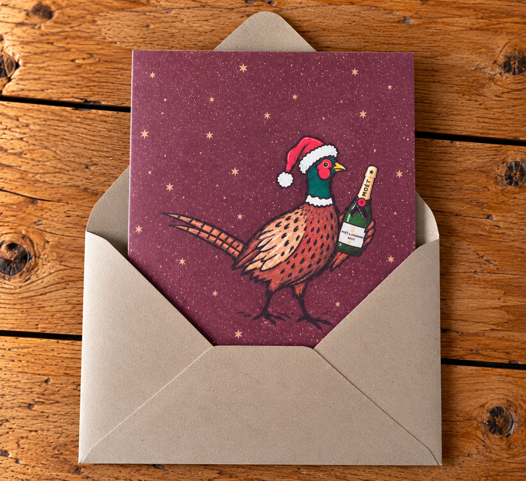 A Christmas-themed greeting card featuring a pheasant holding a bottle of champagne under a Santa hat, displayed inside an open kraft paper envelope on a rustic wooden table.