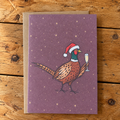 A rectangular greeting card with a purple background and an illustration of a pheasant wearing a Santa hat and holding a champagne flute, placed on a brown kraft envelope over a rustic wooden surface.