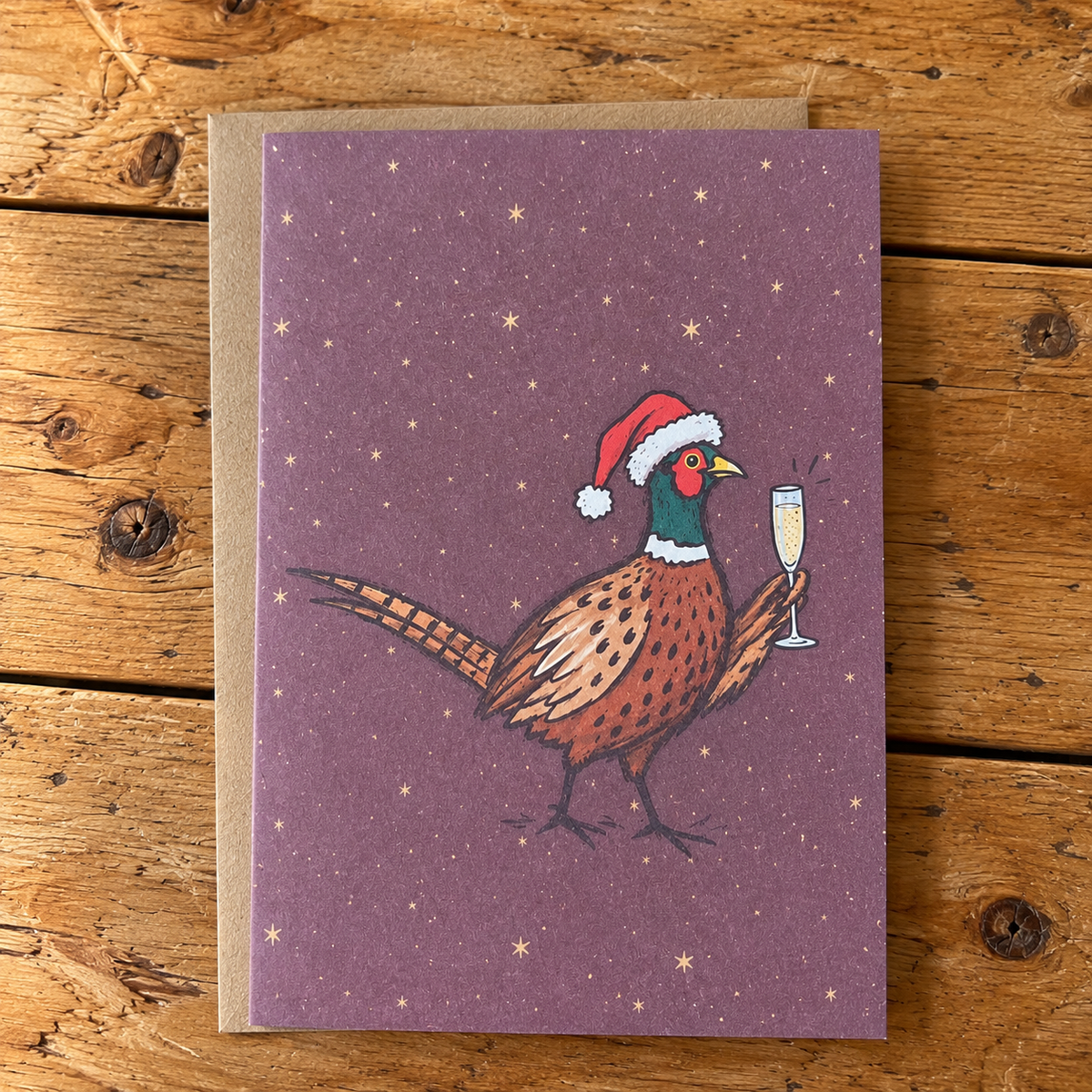 A rectangular greeting card with a purple background and an illustration of a pheasant wearing a Santa hat and holding a champagne flute, placed on a brown kraft envelope over a rustic wooden surface.