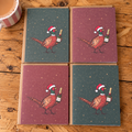 Four square greeting cards arranged in a 2x2 grid on a rustic wooden table. Each card features an illustration of a pheasant wearing a Santa hat and holding a champagne bottle, with two cards in dark teal and two in dusty red

