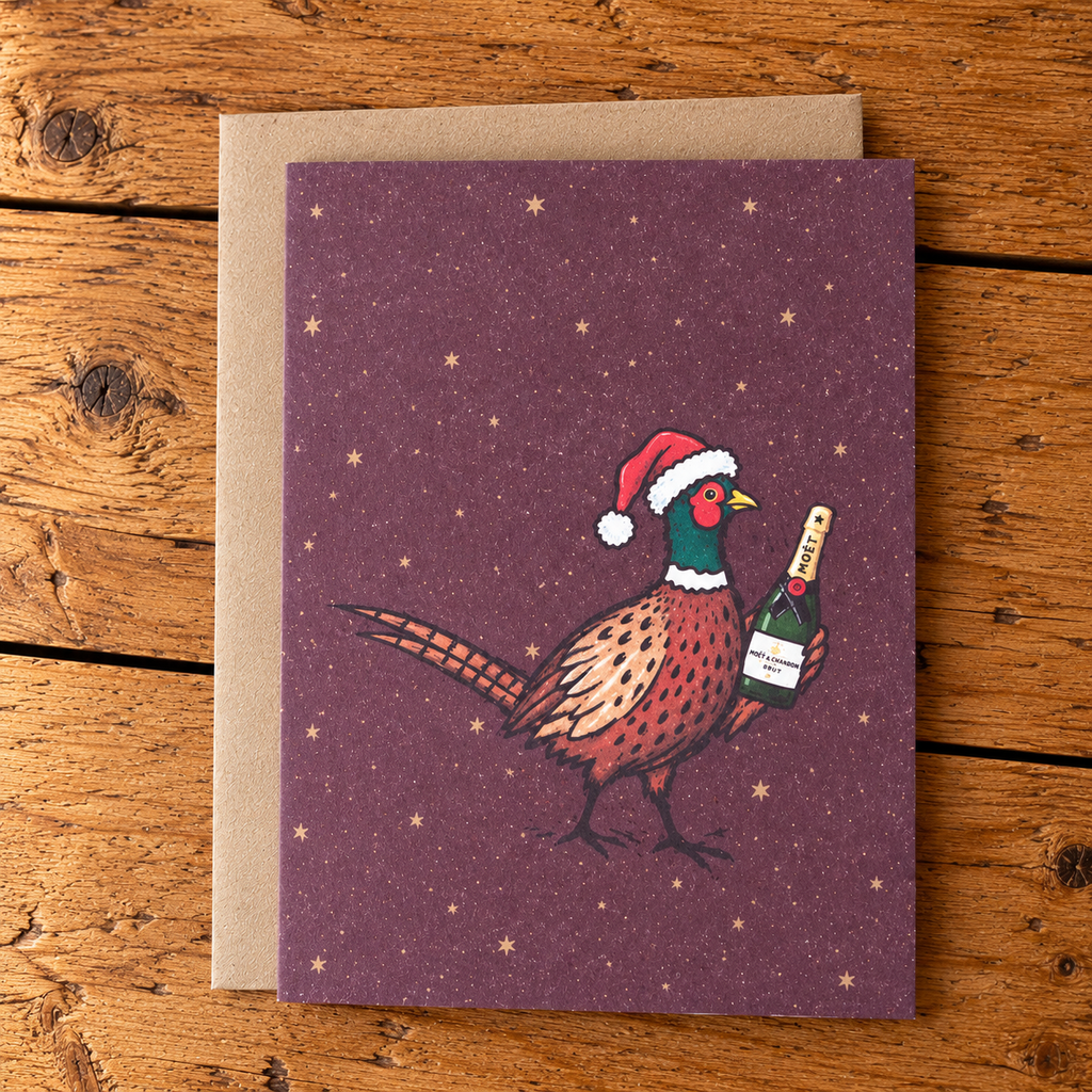 A Christmas-themed greeting card featuring an illustration of a pheasant wearing a Santa hat and holding a champagne bottle, set against a starry purple background. The card is shown with a brown kraft envelope on a rustic wooden surface.