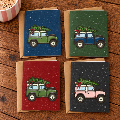 Set of four Christmas greeting cards from The Pink Pheasant featuring illustrated vintage Land Rover-style vehicles carrying Christmas trees tied to their roofs with red ribbons. Designs in green, blue and pink with navy, forest green, red and charcoal snowy backgrounds. Displayed flat with individual kraft envelopes on a rustic wooden table beside a pink striped mug.