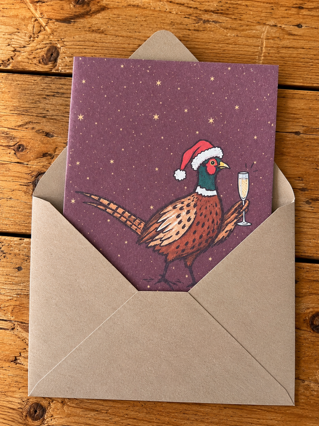 A Christmas-themed greeting card featuring an illustration of a pheasant wearing a Santa hat and holding a champagne glass, set against a dark purple starry background. The card is partially tucked into a brown kraft paper envelope, displayed on a rustic wooden surface.