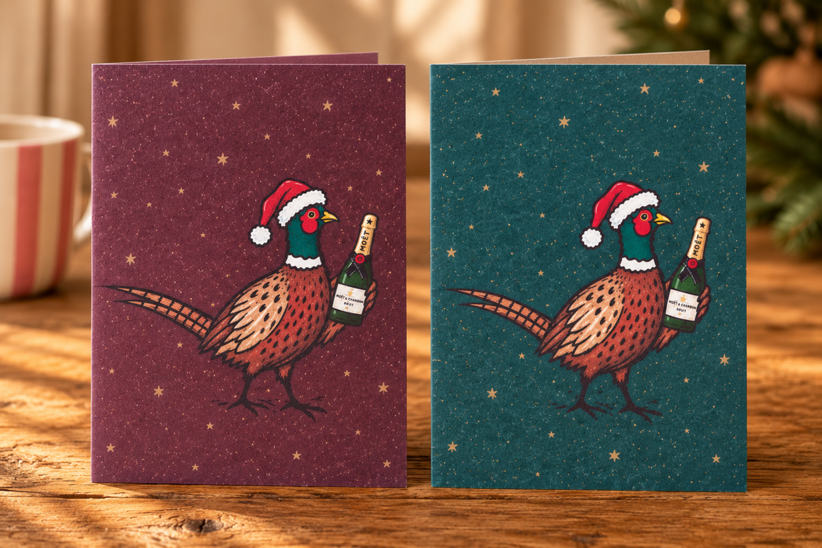 Two Christmas greeting cards displayed side by side on a wooden table. The left card has a dark burgundy background with gold stars and an illustration of a pheasant wearing a Santa hat and holding a champagne bottle.