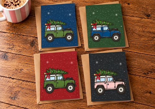 Four Christmas-themed greeting cards arranged on a rustic wooden table, each showing a small jeep carrying a Christmas tree, with kraft paper envelopes. A mug is partially visible in the top-left corner.