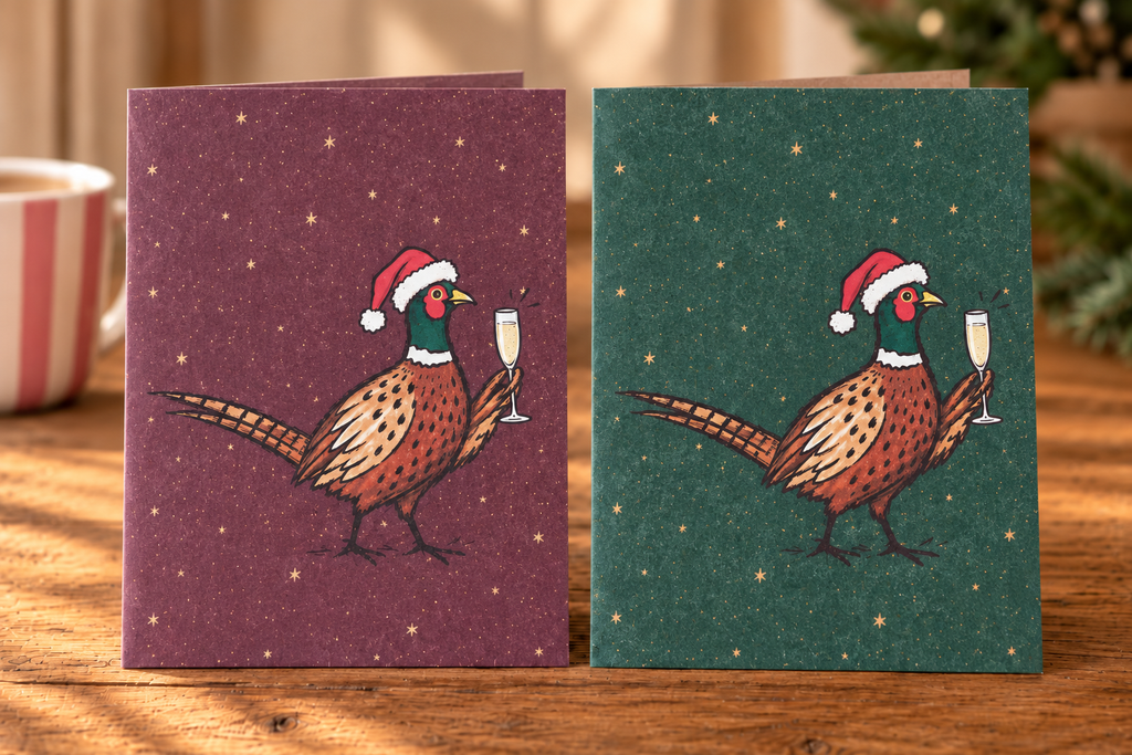 Two Christmas-themed greeting cards displayed side by side on a wooden table. The left card has a dark plum background with a pheasant wearing a Santa hat and holding a glass of champagne, and the right card has a dark green background with the same pheasant illustration.