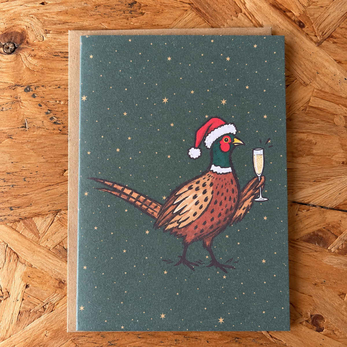 A rectangular greeting card featuring an illustrated pheasant wearing a Santa hat and holding a champagne flute, set against a dark starry background. The card is placed on a brown kraft envelope over a wooden surface.
