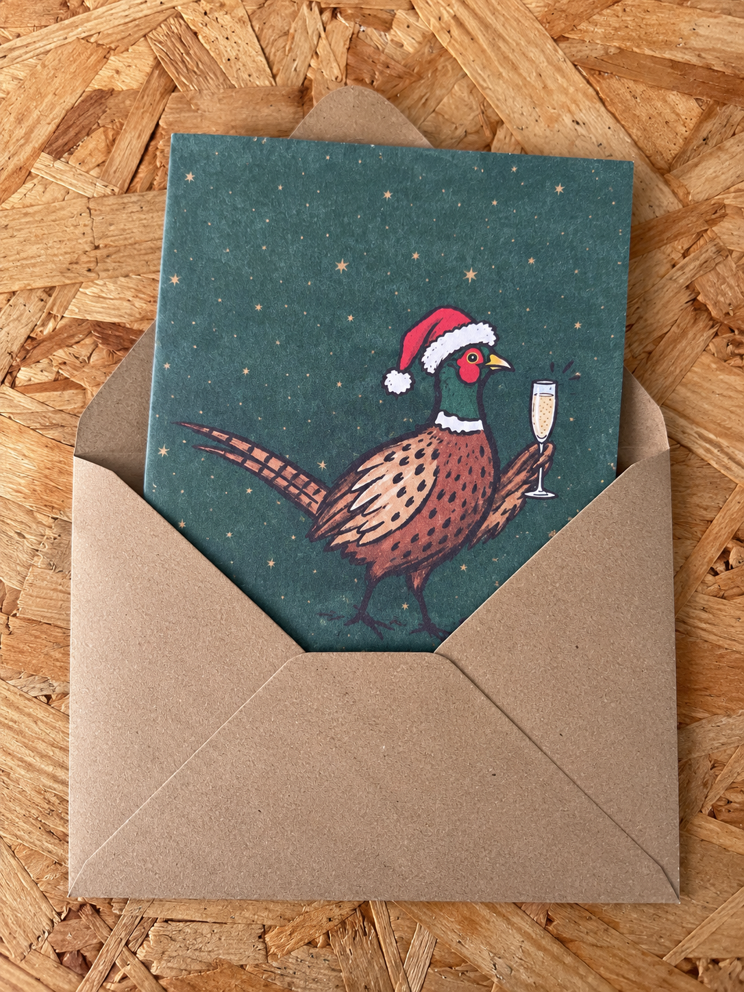 A square greeting card with a brown pheasant wearing a red Santa hat and holding a champagne glass, set against a dark teal starry background. The card is partially tucked into a brown kraft paper envelope, placed on a light brown wood OSB board background.