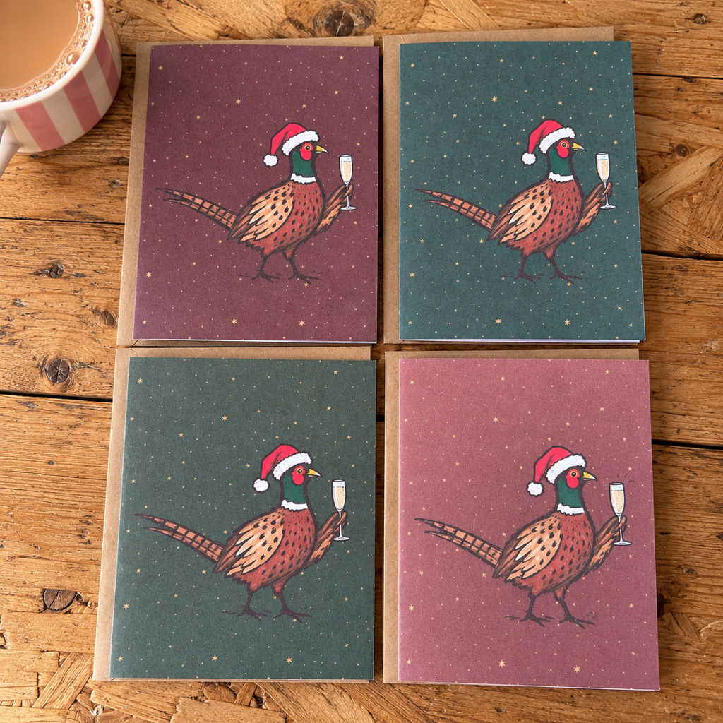 Four square greeting cards arranged in a 2x2 grid on a rustic wooden table. Each card features an illustrated pheasant wearing a Santa hat and holding a champagne glass, with two cards in muted plum pink and two in dark teal.