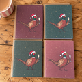 Four square greeting cards arranged in a 2x2 grid on a rustic wooden table. Each card features an illustrated pheasant wearing a Santa hat and holding a champagne glass, with two cards in muted plum pink and two in dark teal.