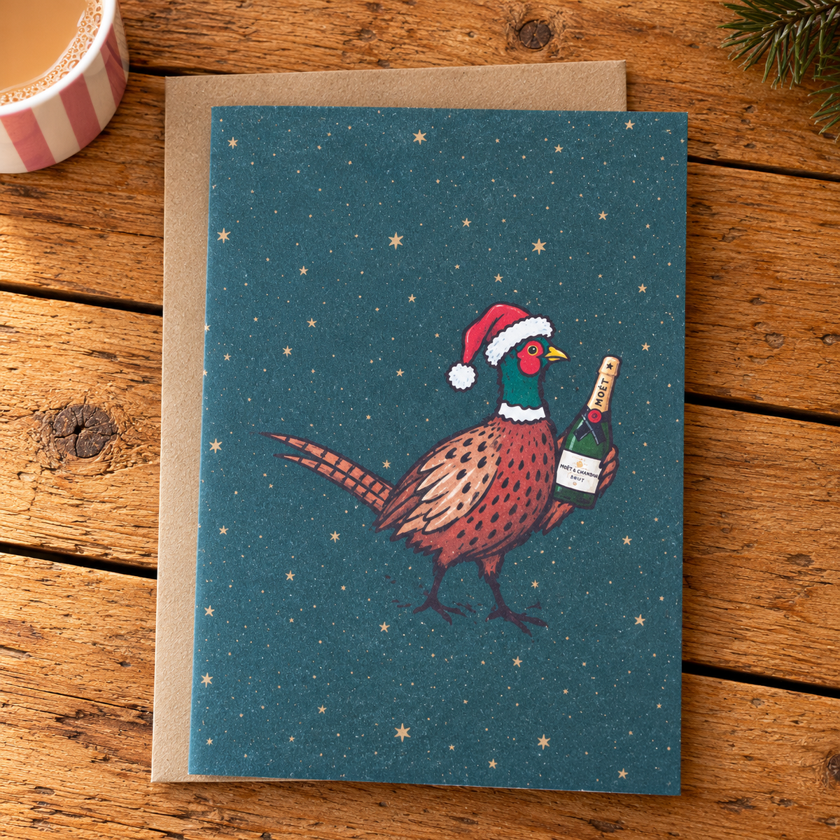 A square greeting card with a brown envelope, featuring an illustration of a pheasant wearing a Santa hat and holding a champagne bottle against a dark teal starry background, styled on a wooden tabletop.